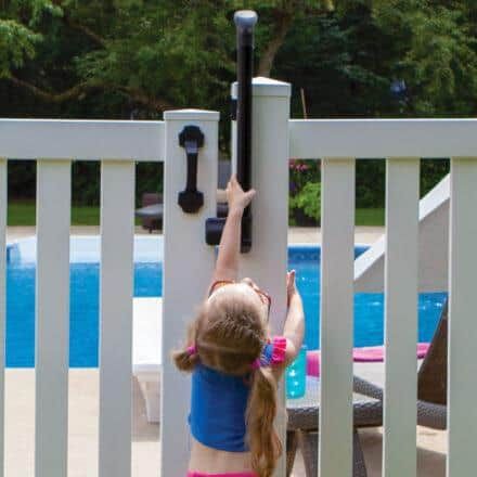 Child reaching up to open a white gate beside a backyard pool with a long black handle