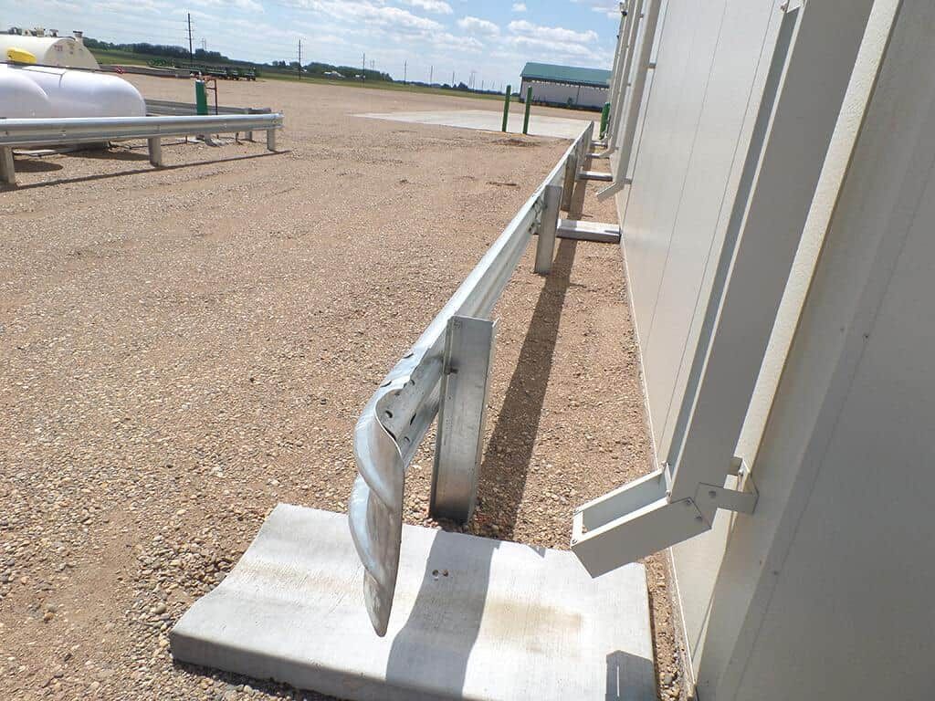 Bent metal guardrail beside a white building on a gravel lot, with a damaged base and bent support