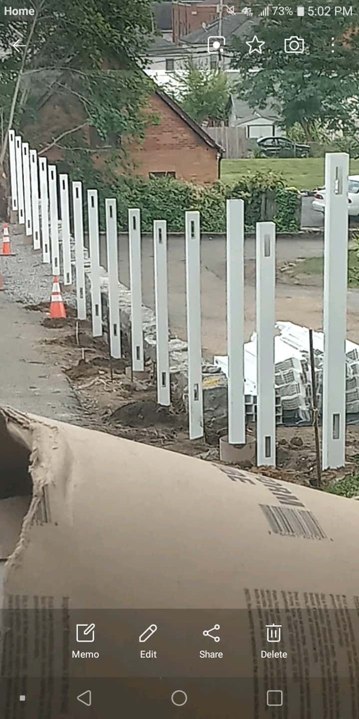 Row of white fence posts along a muddy roadside beside brick houses and trees