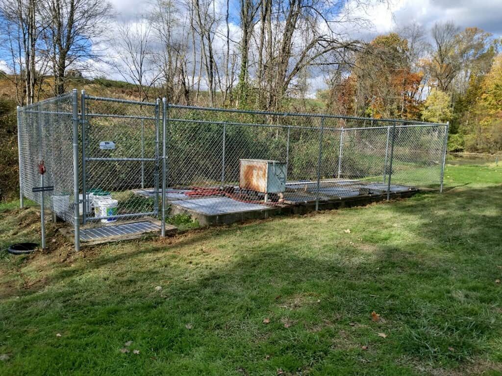 Chain-link animal pen with raised platforms and shelter in a grassy yard under trees