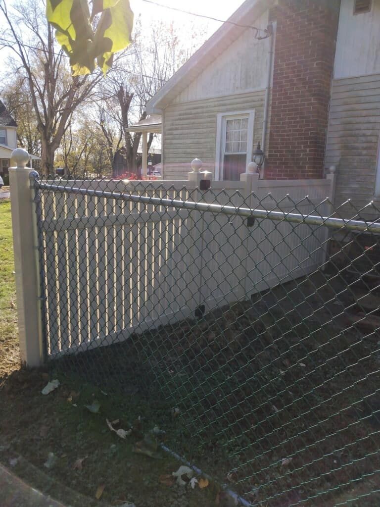 Chain-link fence beside a beige house with a brick wall and yard in autumn sunlight