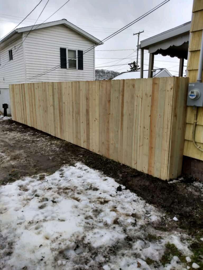 New wooden privacy fence beside a house on a snowy, muddy backyard lot