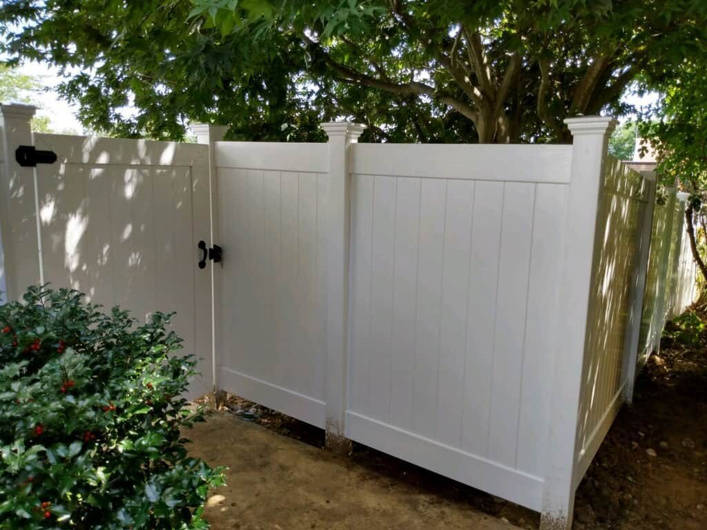White backyard gate and fence with black hardware beside a tree and shrubs