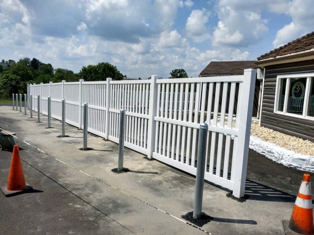 White picket fence panels along a concrete walkway beside a building, with orange cones and blue sky