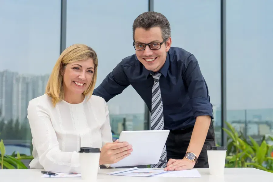 Two coworkers smiling over a document in a bright office, with coffee cups on the table