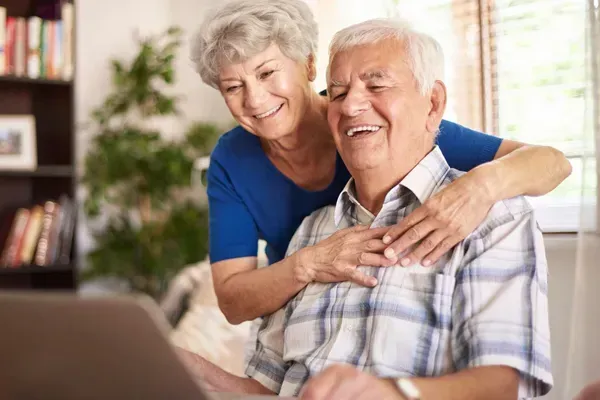Smiling older couple at a laptop, woman hugging man from behind in a bright room