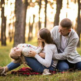 Family sitting on grass in a sunlit park, smiling and cuddling with a child lying across their laps
