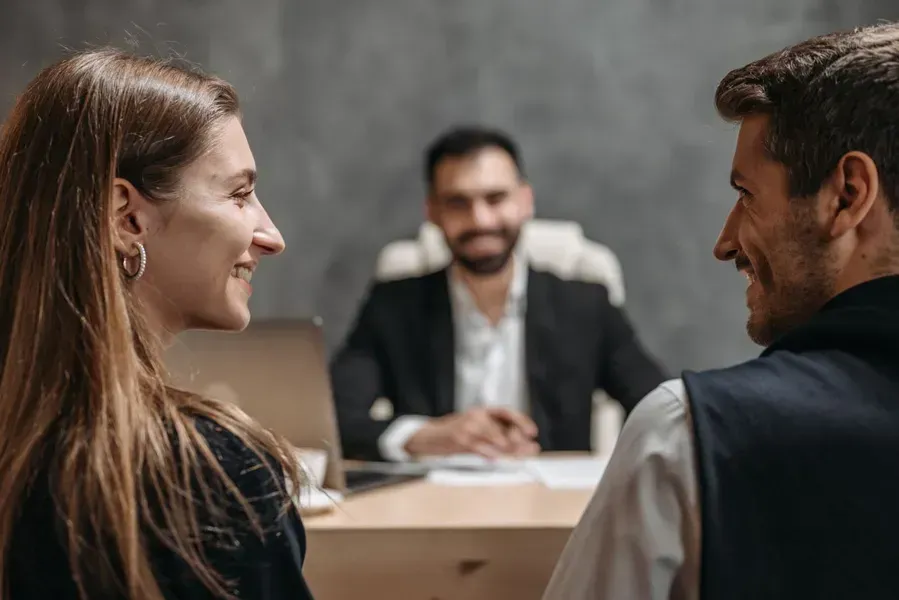 Two people smiling across a desk in a meeting room, with a third person seated in the background.