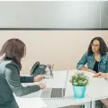 Two people in a meeting across a white desk, with a laptop, phone, and small potted plant.