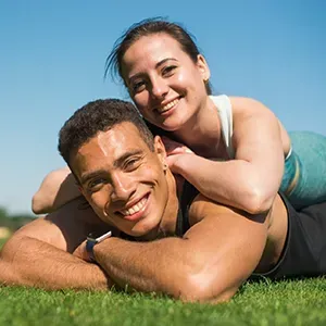 Smiling couple lying on green grass, with one person resting on the other's back under a blue sky