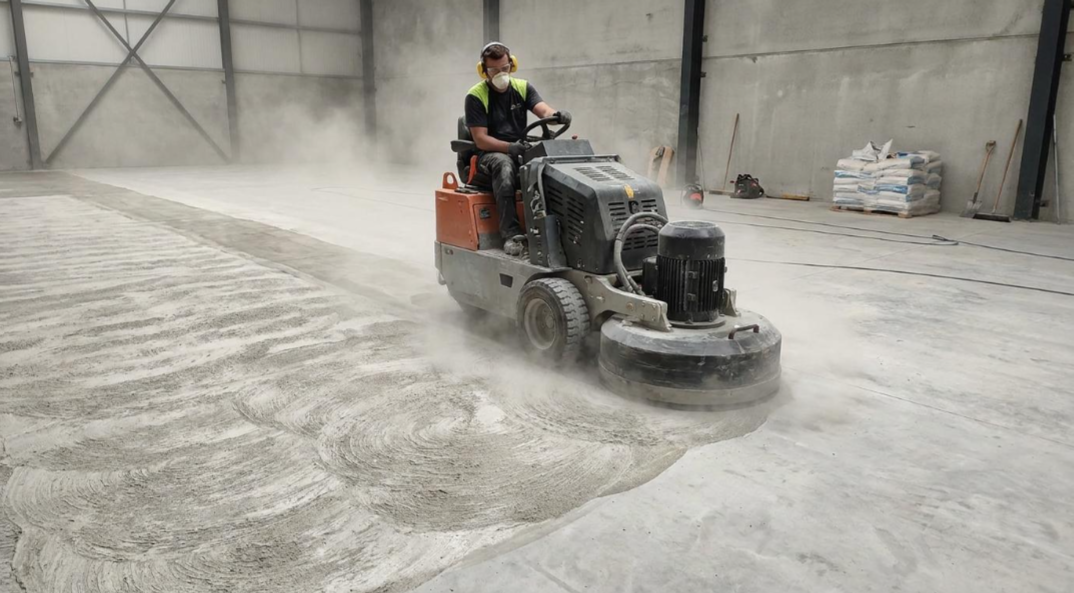Worker operating a floor scrubber in a dusty industrial warehouse, spraying water across concrete.