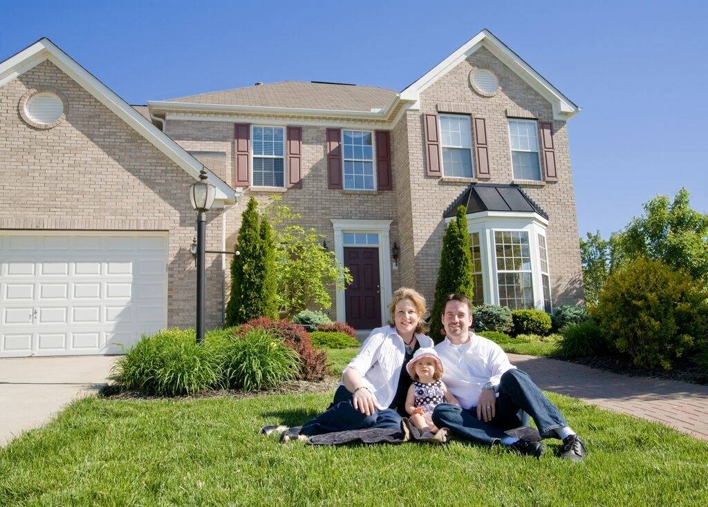 Par une journée ensoleillée, une famille est assise sur la pelouse devant une maison de banlieue en briques de deux étages.