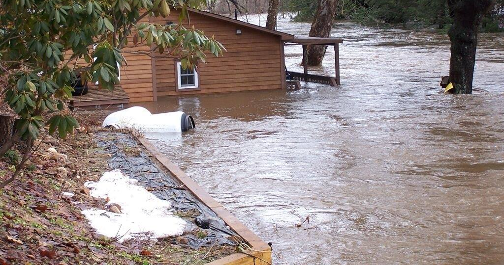 Une maison en bois est partiellement submergée par les eaux de crue, avec un objet blanc partiellement enfoui