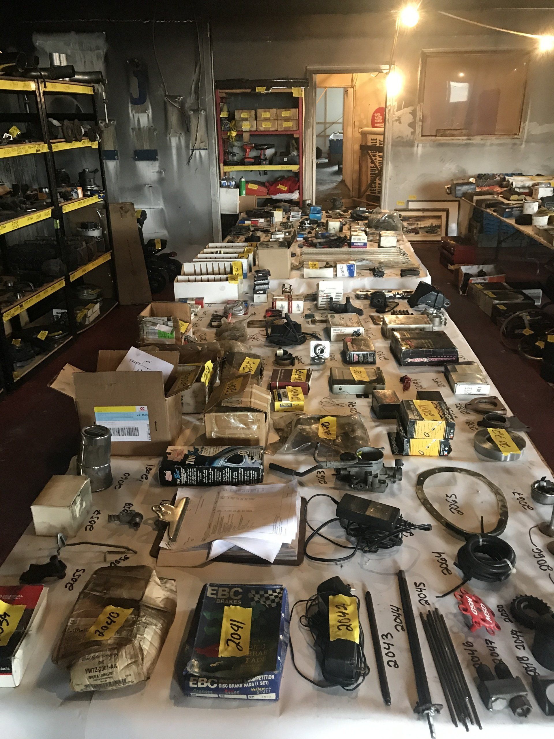 Un établi encombré dans un atelier, où sont étalées diverses pièces et fournitures automobiles triées.