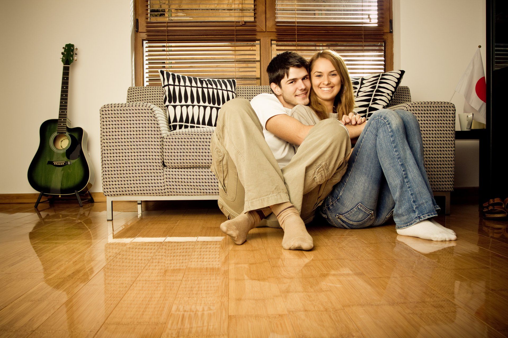 Couple sitting on floor, embracing, near patterned couch and guitar; happy smiles.