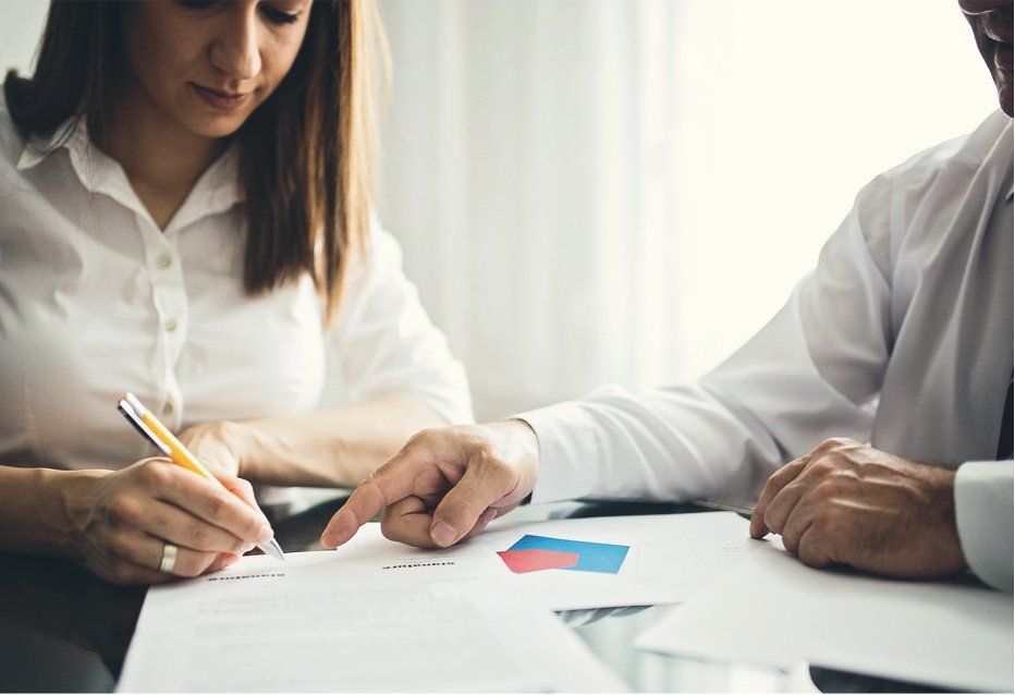 Une femme signe un document sous le regard d'un homme. Tous deux portent des chemises blanches et se trouvent dans un bureau.