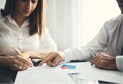 Une femme signe un document sous le regard d'un homme. Tous deux portent des chemises blanches et se trouvent dans un bureau.