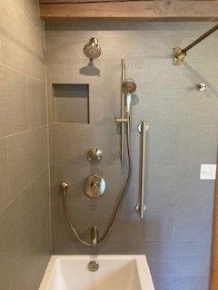 Bathroom shower with gray tiled walls, showerheads, a handrail, and a tub.