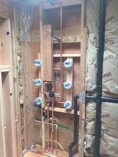 Copper plumbing and black drain pipes installed in a wooden bathroom wall. Insulation surrounds.