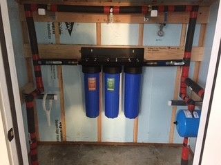 Water filtration system in a utility room. Blue filter housings mounted on wooden frame, with piping and pressure tank.