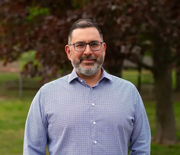 Man in glasses and checkered shirt standing outdoors with trees in the background