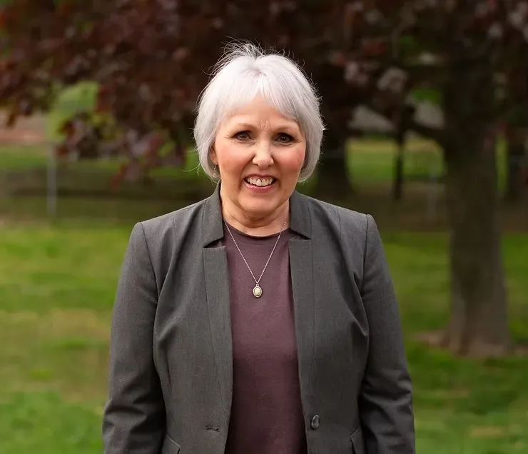 Smiling woman with short white hair in a gray blazer standing outdoors in a park