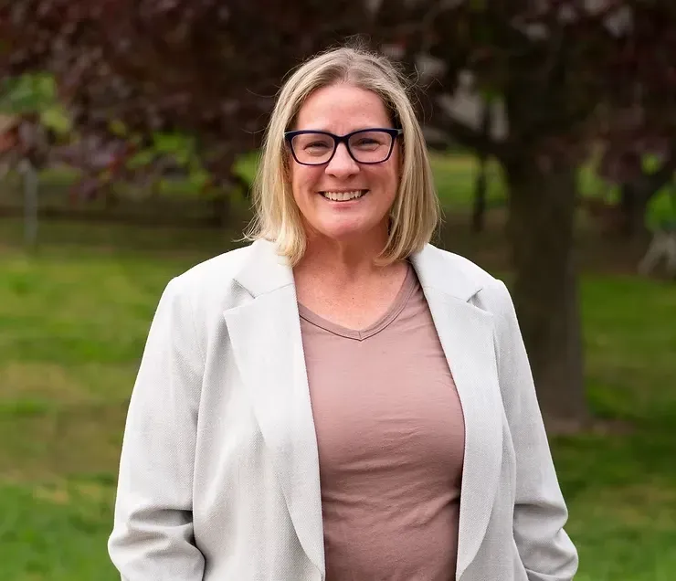 Smiling person in glasses and white blazer standing outdoors in a park.