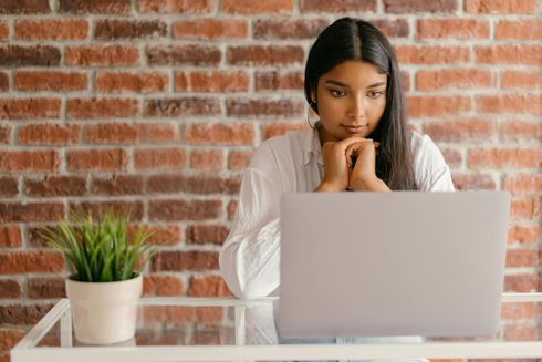 Person resting chin on hands behind a laptop at a desk, with a brick wall and potted plant in the background.