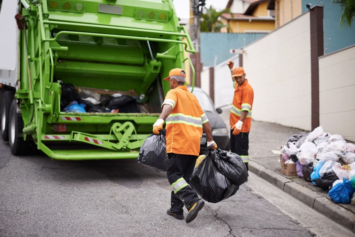 Garbage collectors in orange uniforms load trash bags into a green garbage truck on a street