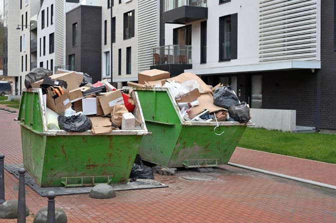 Two overflowing green dumpsters full of cardboard and trash beside modern apartment buildings.