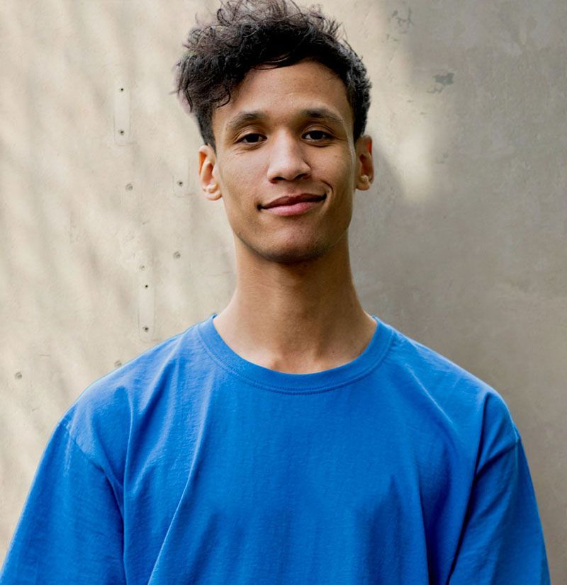 Man with curly dark hair smiles, wearing a blue t-shirt, standing in front of a light-colored wall.