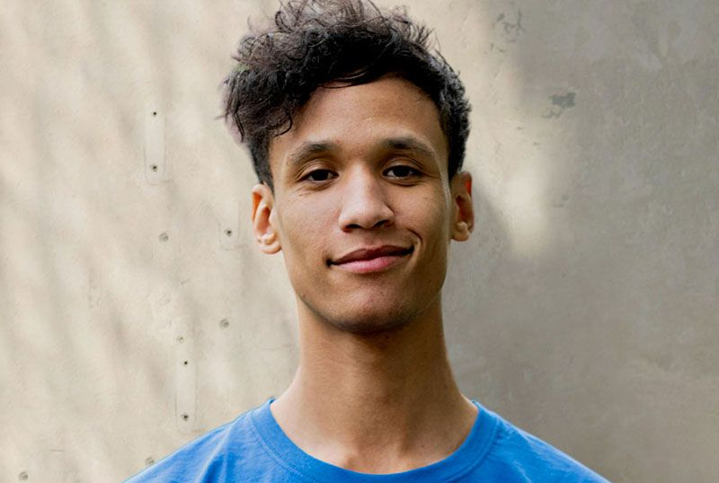 Man wearing blue shirt smiling in front of a concrete wall.