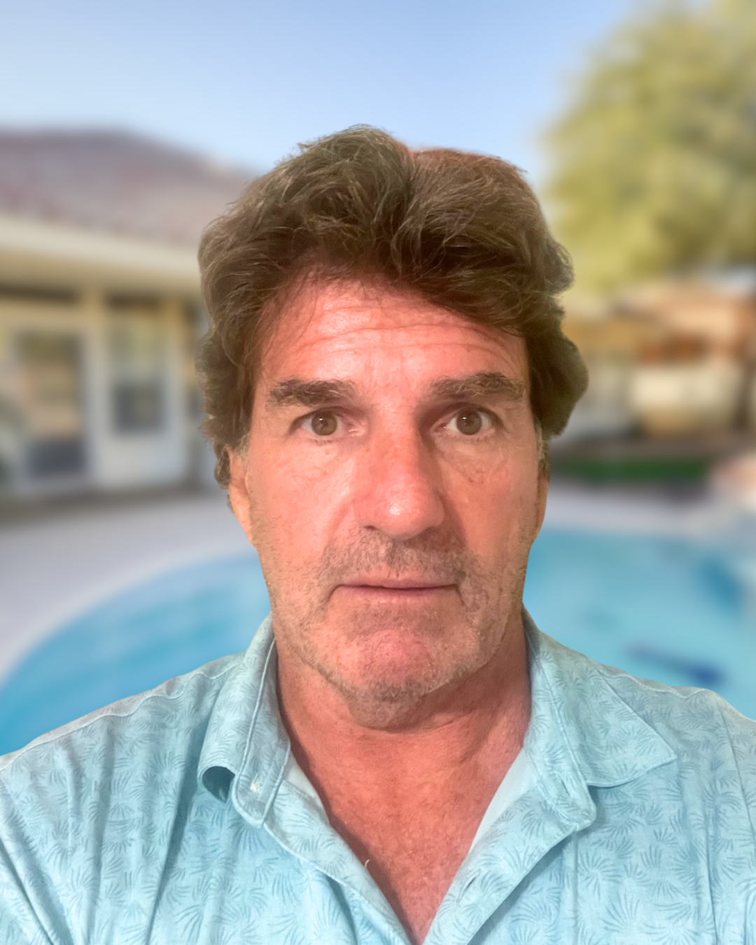 Man in light blue shirt, looking directly at the camera with a swimming pool blurred in the background.