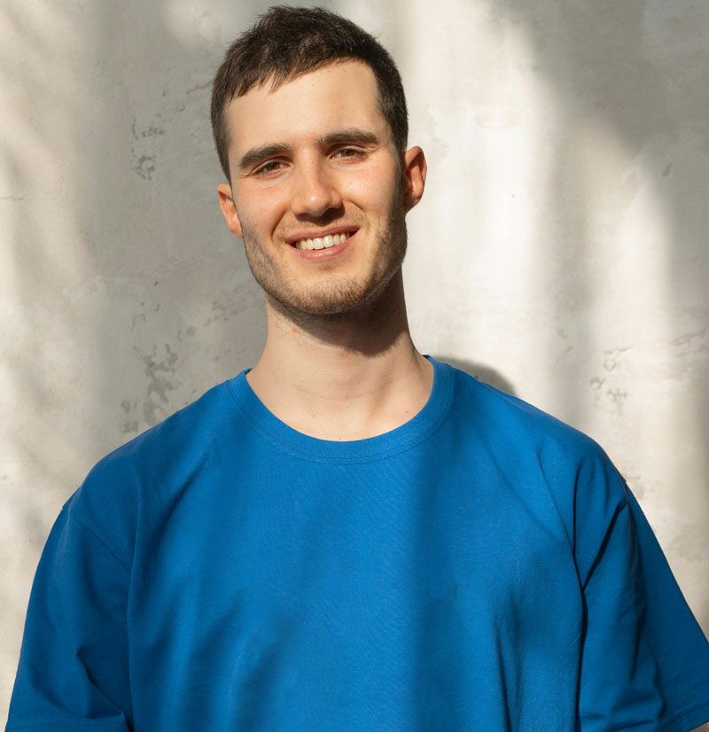 Man with short brown hair smiling, wearing a blue shirt against a light-colored wall with shadow.