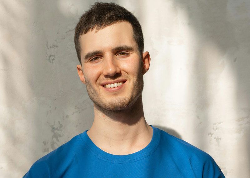 Man in blue shirt smiles in front of a textured light wall, with shadows.
