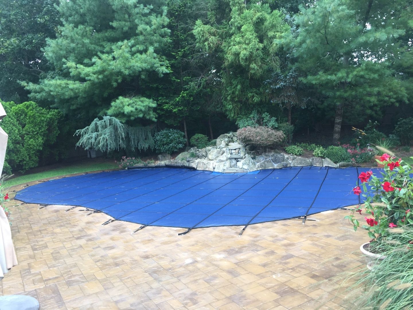 Blue pool cover over a pool with a waterfall feature, surrounded by trees and a paved patio.