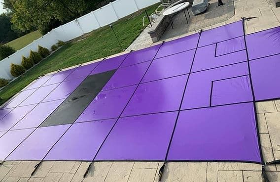 Purple pool cover on a stone patio, covering a rectangular pool. Green lawn and white fence in background.
