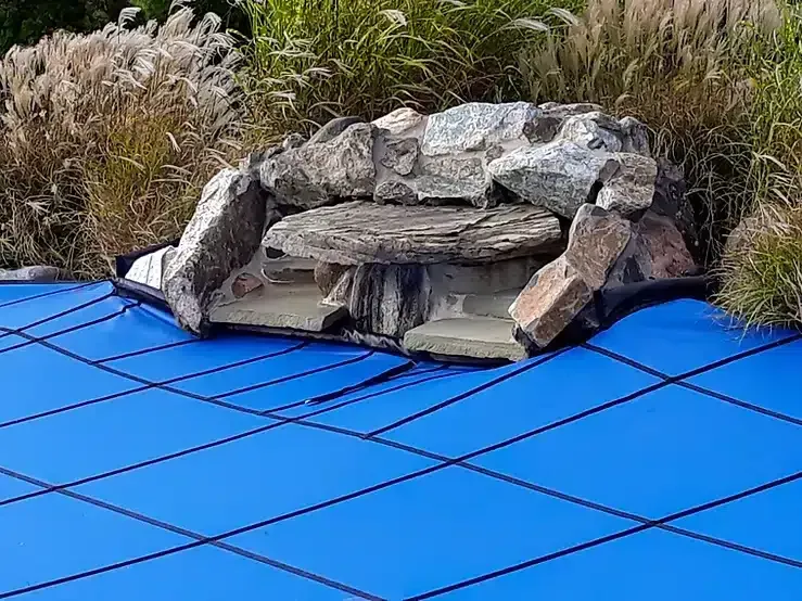 Blue pool cover with a rock waterfall feature in a backyard setting.