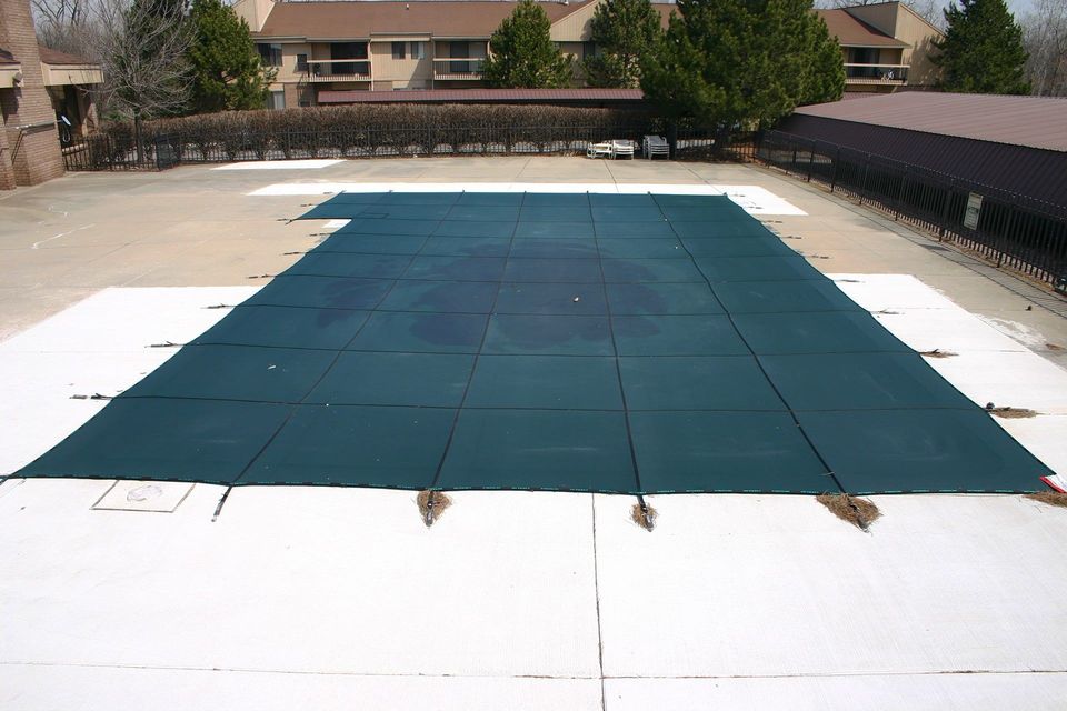 Dark green pool cover on white concrete, surrounded by trees and buildings.
