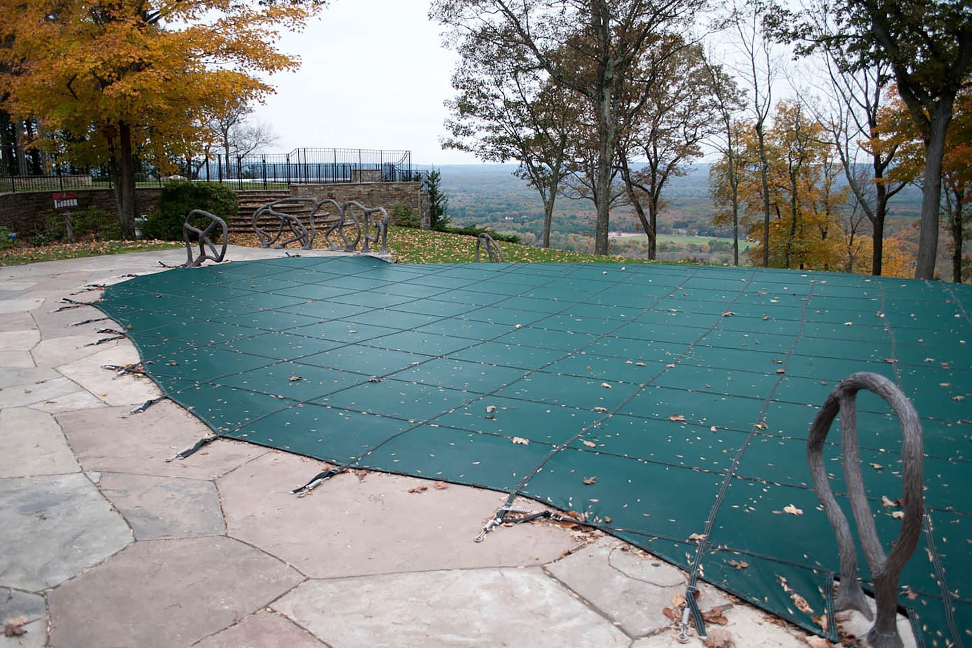 Green pool cover on a stone patio, autumn trees in the background, overcast sky.