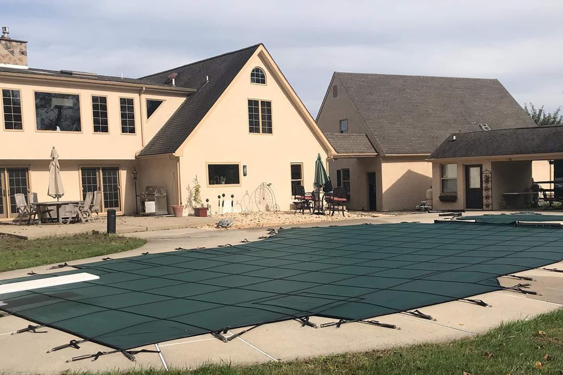 House with pool covered in a green tarp. Beige exterior, brown roof, and patio furniture visible.