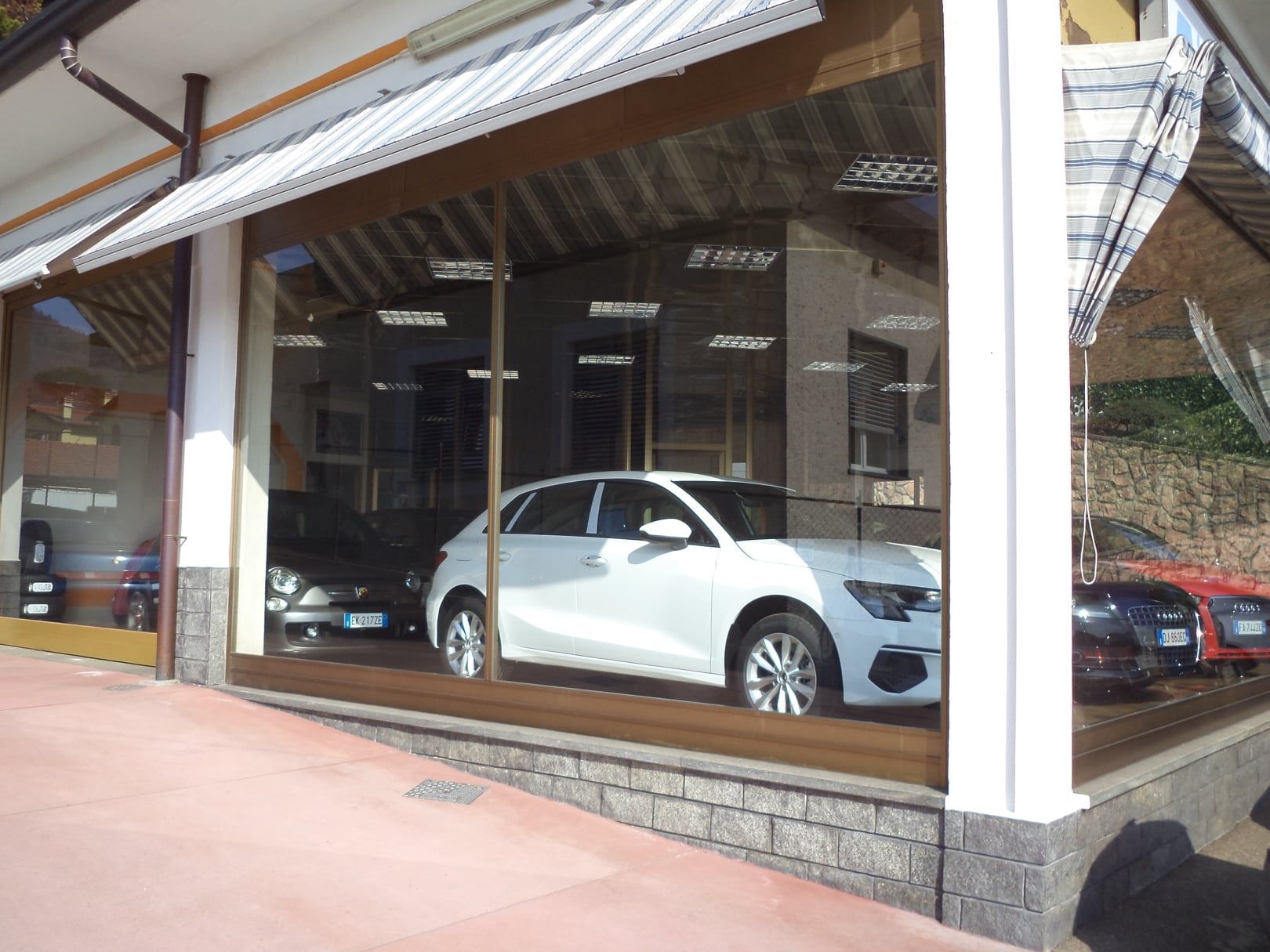 Un SUV bianco esposto in uno showroom con pareti di vetro e un'auto rossa parcheggiata accanto.