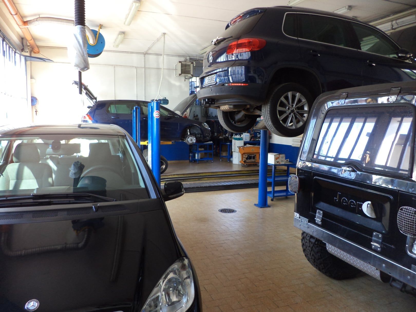 Officina meccanica con un SUV nero sollevato su un ponte blu e auto parcheggiate in primo piano.