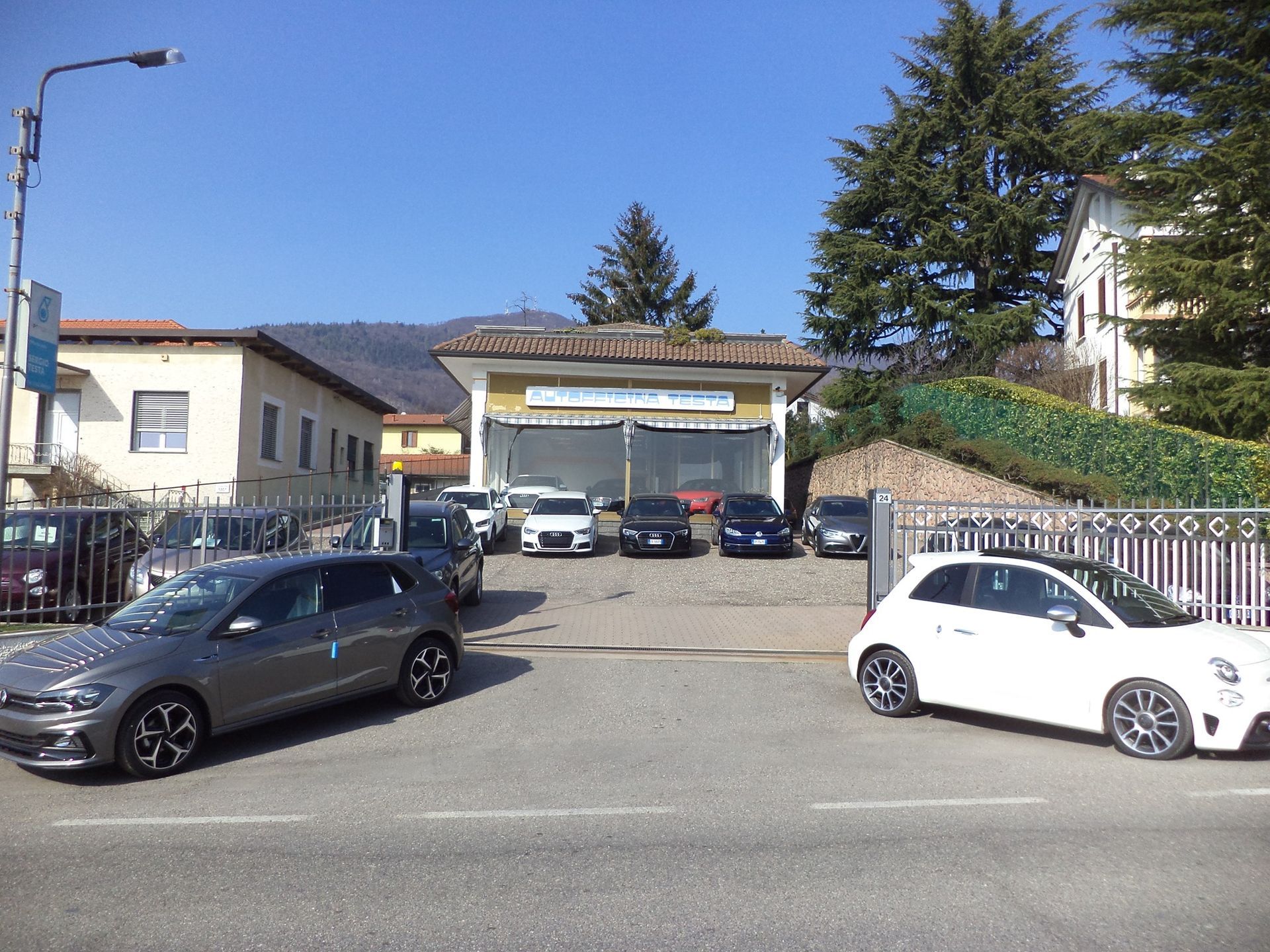 Vialetto di accesso a un'abitazione con diverse auto parcheggiate sotto una tettoia, accanto alle case in una giornata di sole.