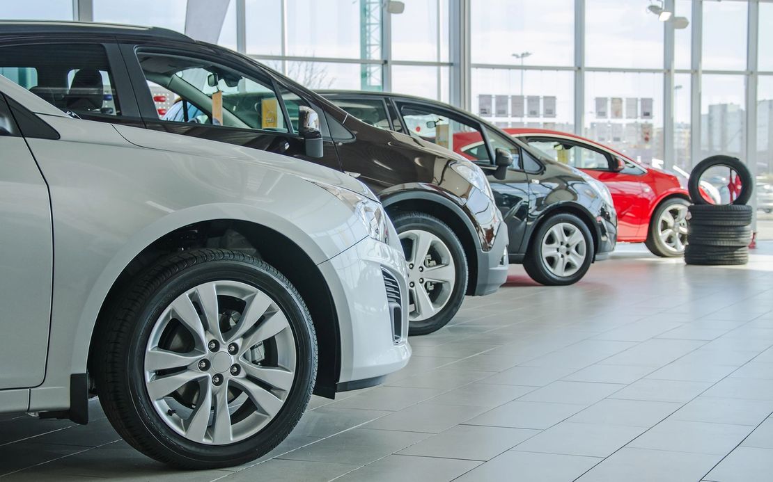 Una fila di auto nuove esposte in uno showroom luminoso, con un'auto bianca in primo piano e auto rosse sullo sfondo.