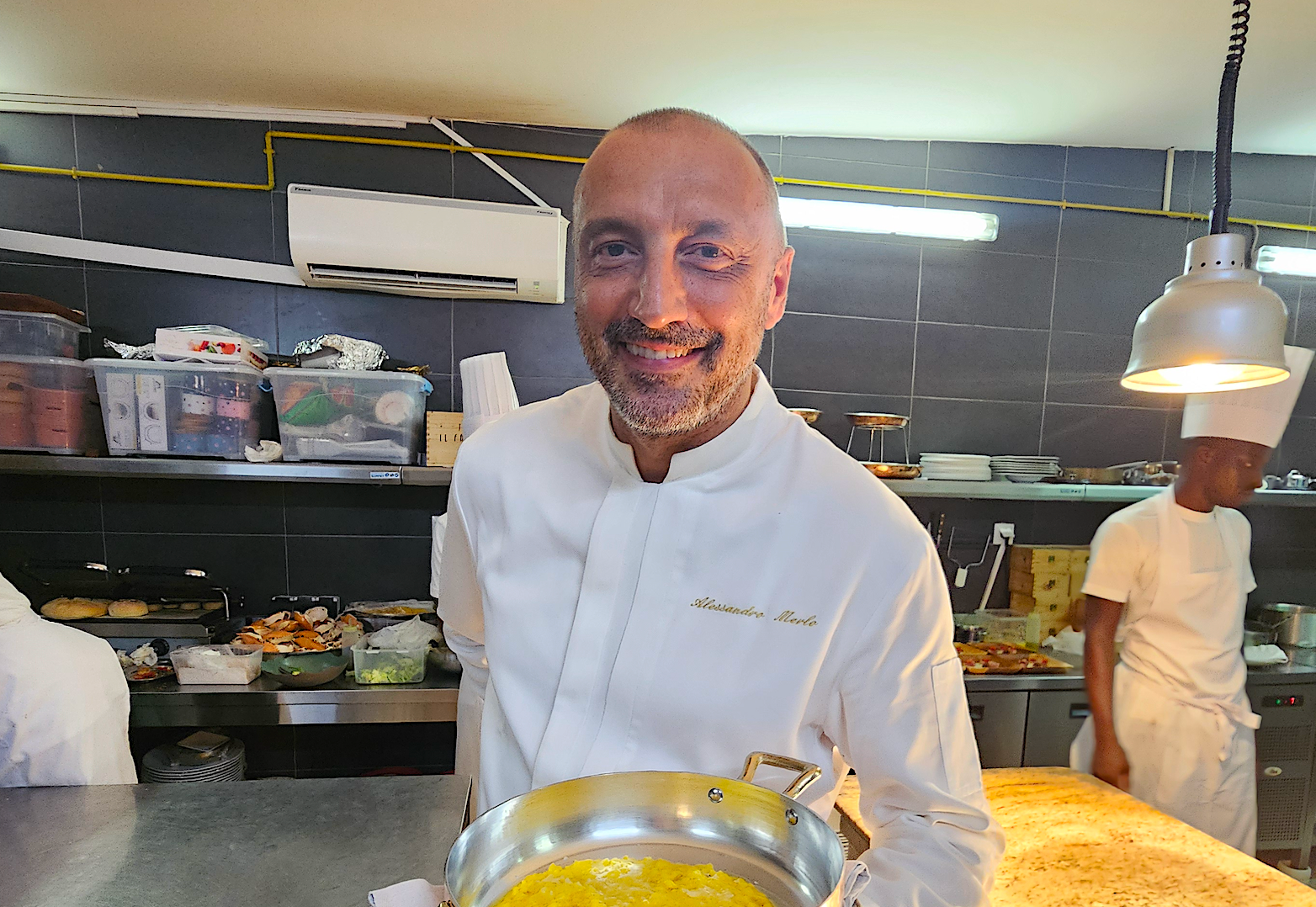 Un chef, souriant, tient un plat dans sa cuisine. On aperçoit en arrière-plan d'autres employés et les zones de préparation.