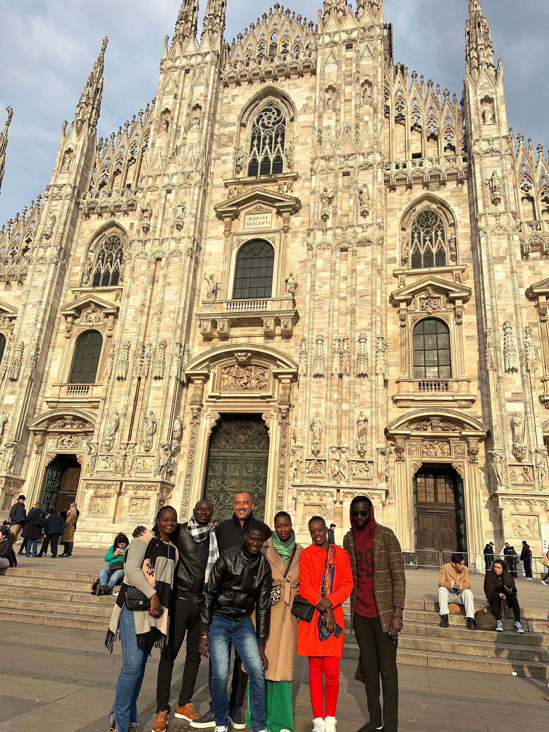 Des photos de groupe sont prises devant la cathédrale de Milan, richement ornée. Des personnes se tiennent debout ou assises.