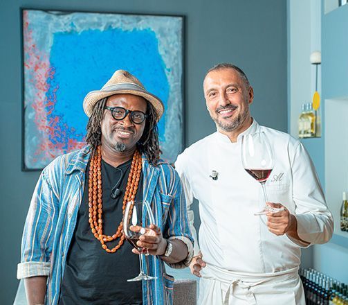 Deux hommes, verres à vin à la main, sourient. L'un porte un chapeau, l'autre une tenue de chef.