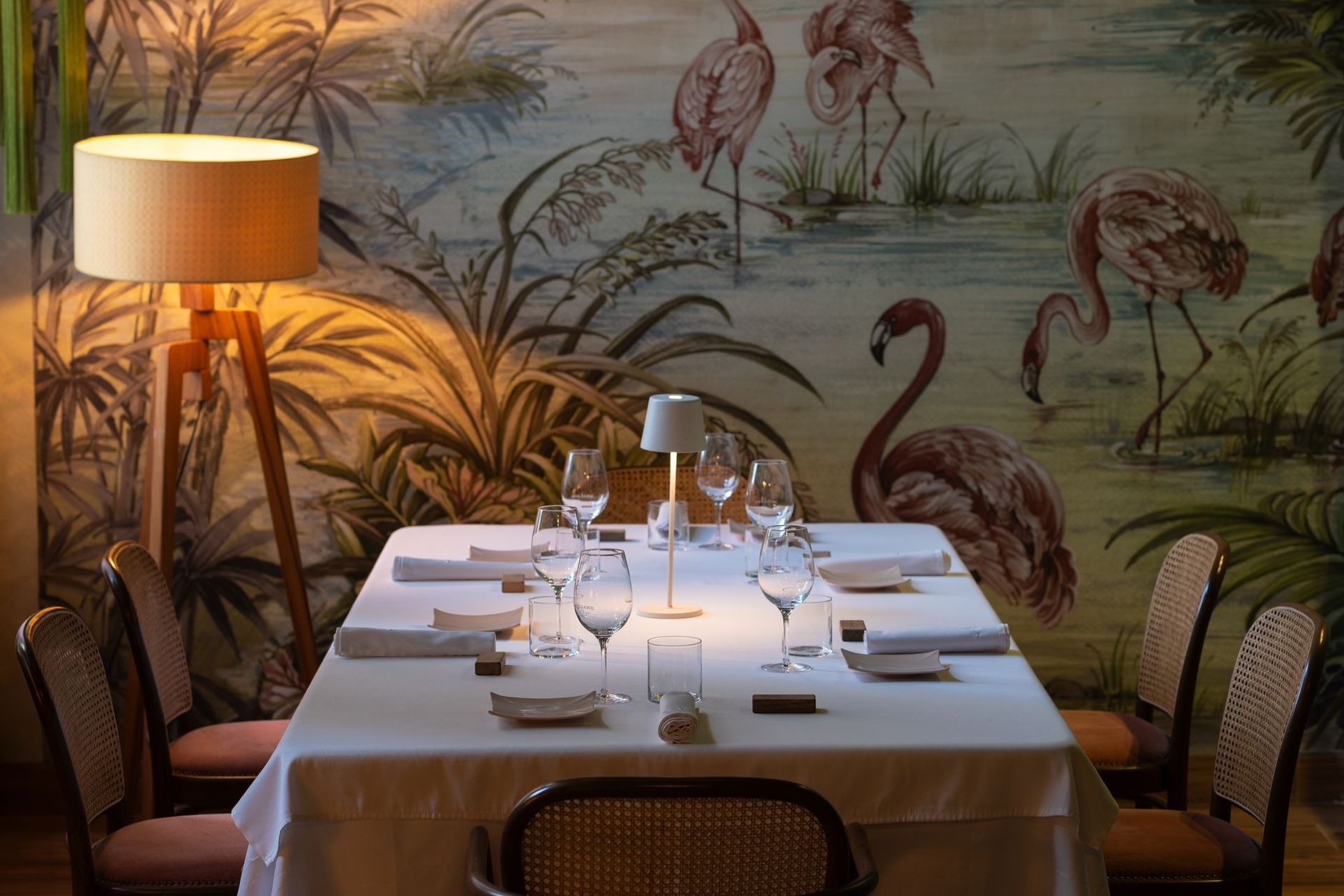 Table à manger dressée pour les invités, devant un mur orné d'une fresque de flamants roses, éclairée par une lampe.