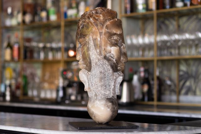 Sculpture à visages multiples, brun clair et gris, sur un comptoir. Derrière, un comptoir flou avec des bouteilles.